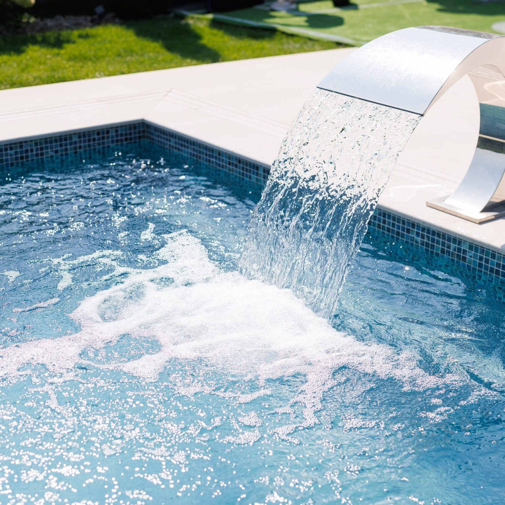 A modern swimming pool with a sleek water feature pouring water into the pool. The water is clear and blue, creating a refreshing atmosphere. The surrounding area is well-maintained with green grass.
